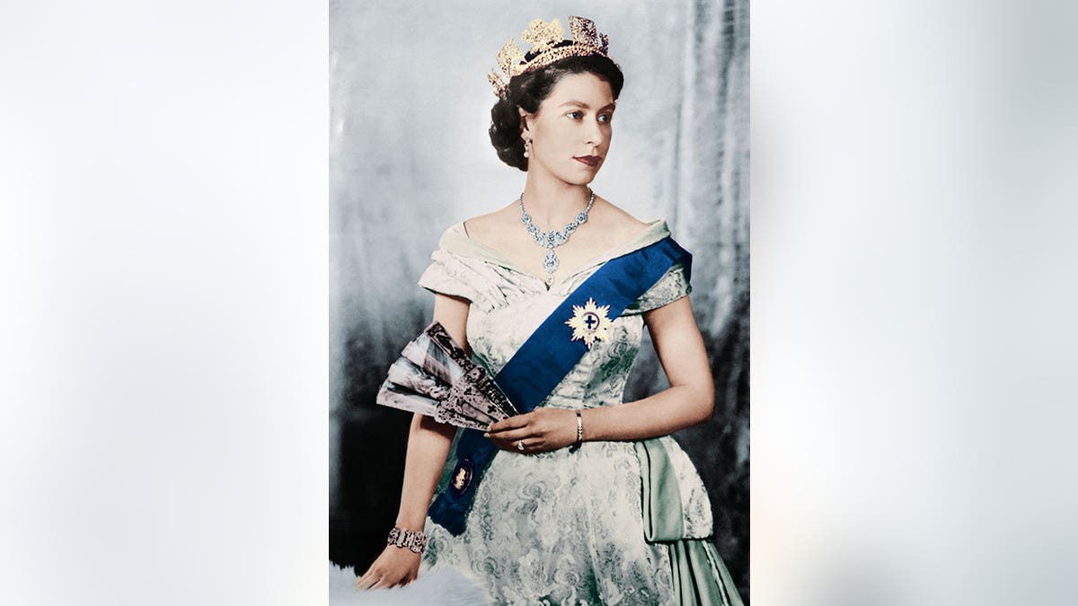Queen Elizabeth wears a white dress, blue sash, and a crown while holding a fan