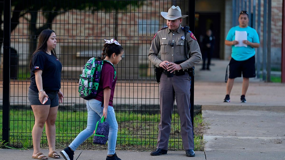 Students with their backpacks walk into school, passing a police officer in uniform