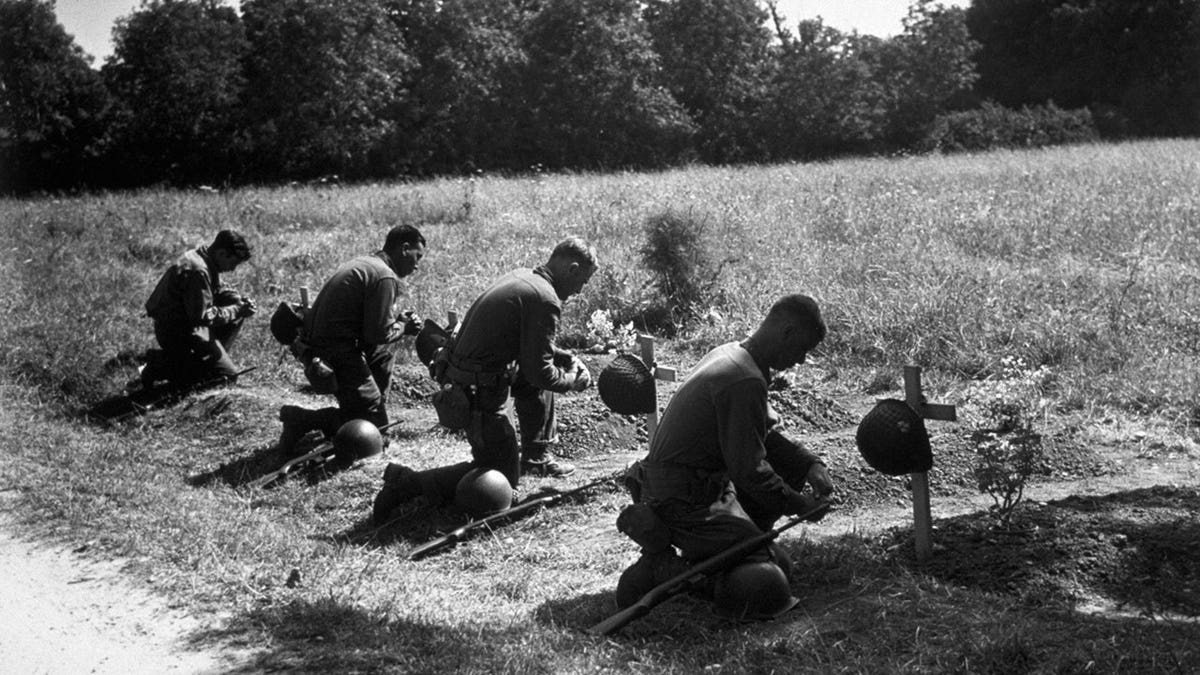 Three soldiers one their knees in front of crosses with helmets laid on top of them in a black and white photo