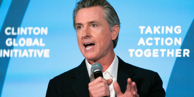 Governor Newsom holding a mic, speaking in front of a blue background