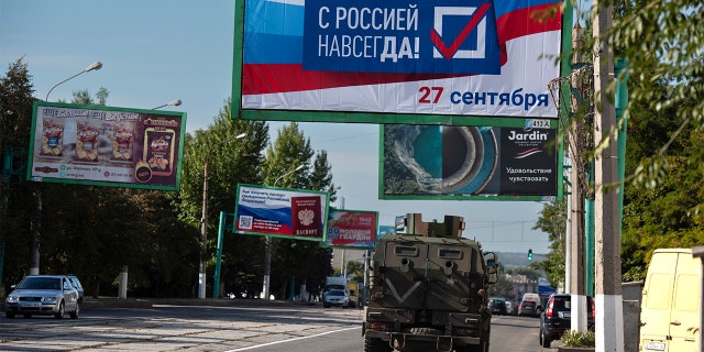 A military vehicle drives along a street with a billboard reading 