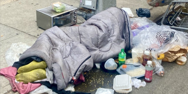 A man sleeps in the rain on the edge of the sidewalk at West 43rd Street in Midtown Manhattan on Sunday, Sept. 11, 2022, just two blocks from Times Square. He had a toaster oven by his side, which he apparently plugged into the base of a city light pole to heat his food.