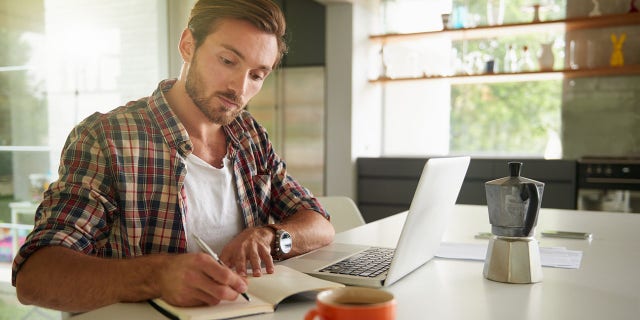 A man writes in his journal while using a laptop at home. Celeste Headlee suggested that people keep track of how they spend their time — and be
