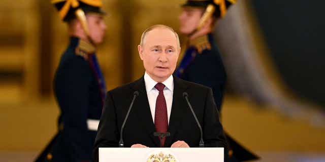 Vladimir Putin delivers an address flanked by men in military uniforms. 