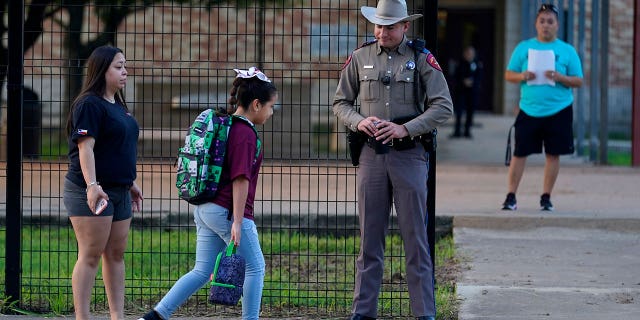 Students with their backpacks walk into school, passing a police officer in uniform