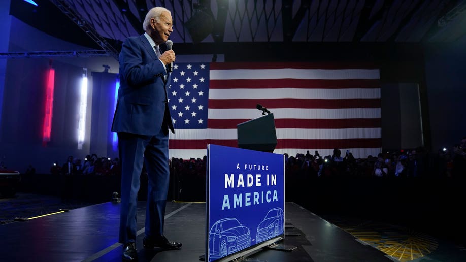 Biden holding a mic, speaking on stage in front of an American flag with a "Made In America" sign in front