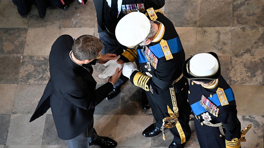 An overhead shot of King Charles having his gloves adjusted by a man in a dark suit during Queen Elizabeth's state funeral.