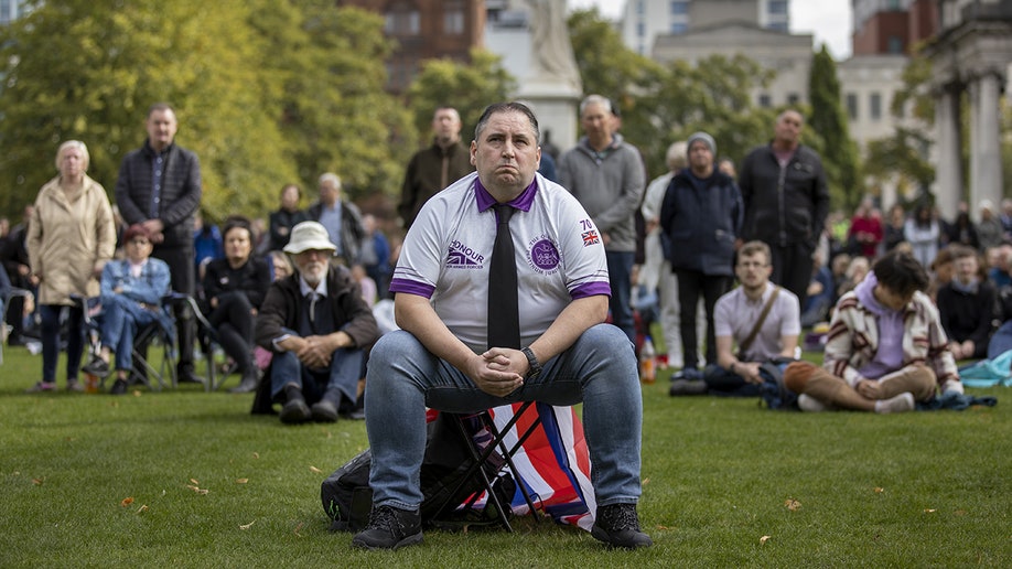 Mourner sits in a park watching Queen Elizabeth's state funeral on a screen.