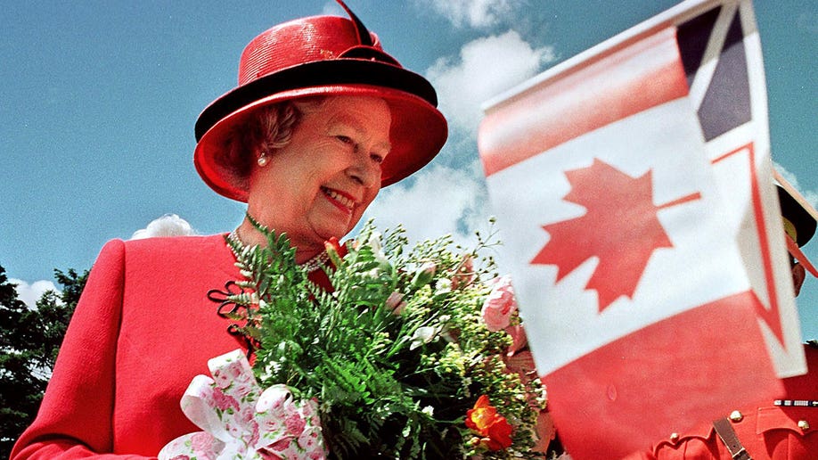 Queen Elizabeth wears a red hat and coat, holds a bouqet of flowers, and smiles while looking at a Canadian flag