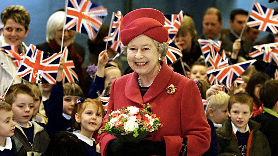 Queen Elizabeth wears a red coat and hat while surrounded by school children holding union jack flags