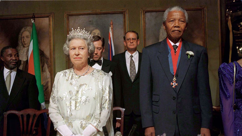 Queen Elizabeth wears a silver dress and a crown while standing beside Nelson Mandela who wears a dark suit