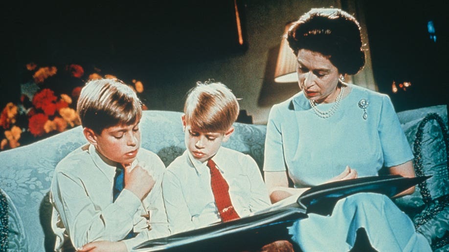 Queen Elizabeth wears a blue dress while holding a photo album; she sits on a couch with her two sons