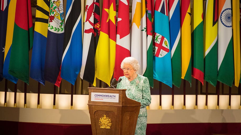 Queen Elizabeth wears a green dress while giving a speech at a podium in front of the Commonwealth's various flags