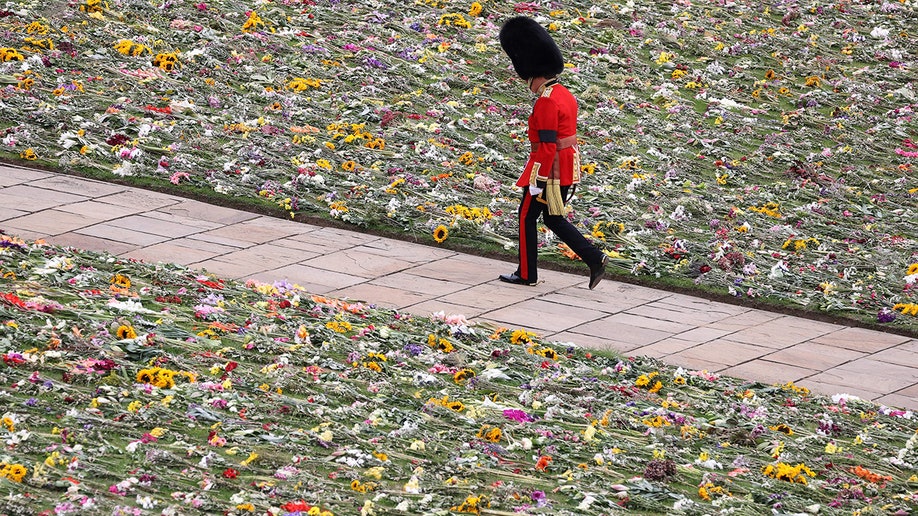 A soldier in a red military uniform walks on a walkway covered with flowers on either side during Queen Elizabeth's state funeral.