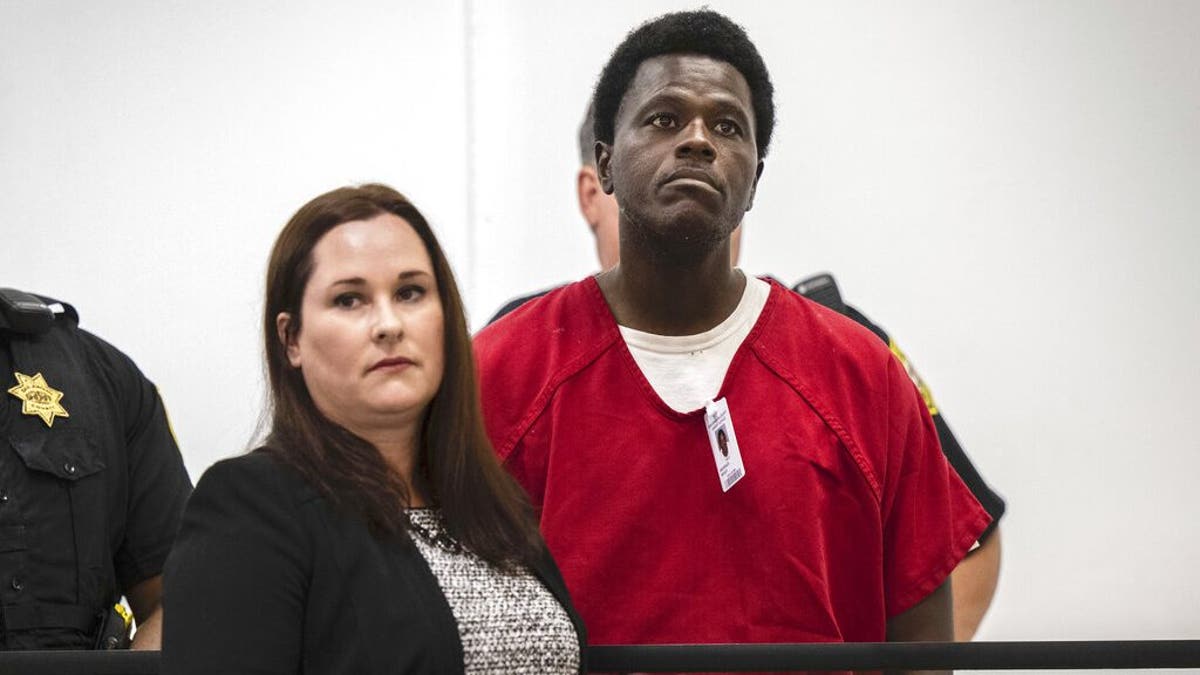 Wesley Brownlee in red prison jumpsuit, standing behind public defender at arraignment