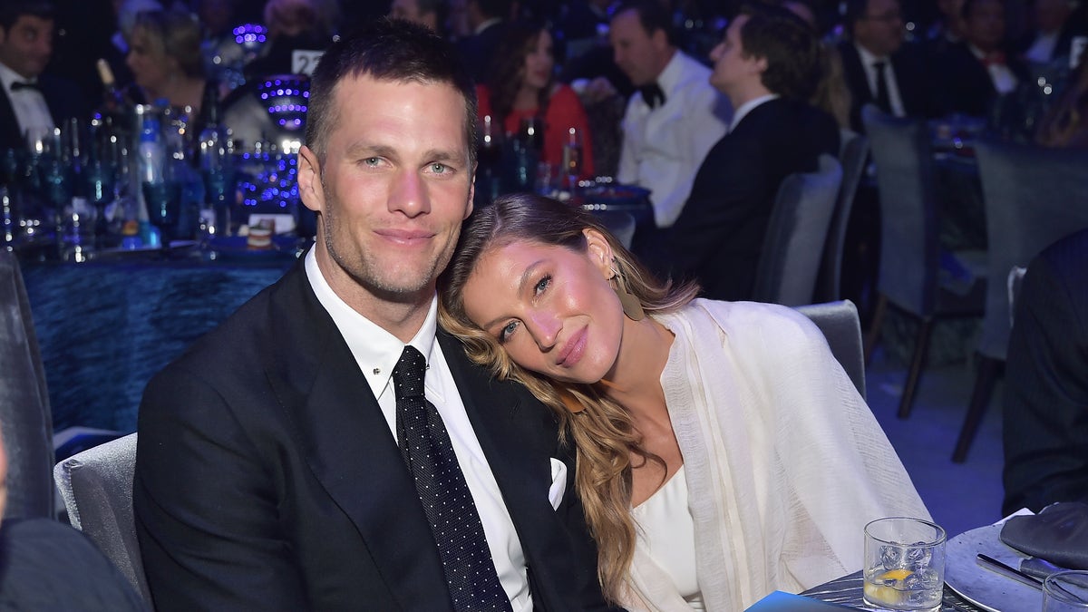 Gisele Bündchen in a white shirt and cream cardigan puts her head on Tom Brady's shoulder in a suit