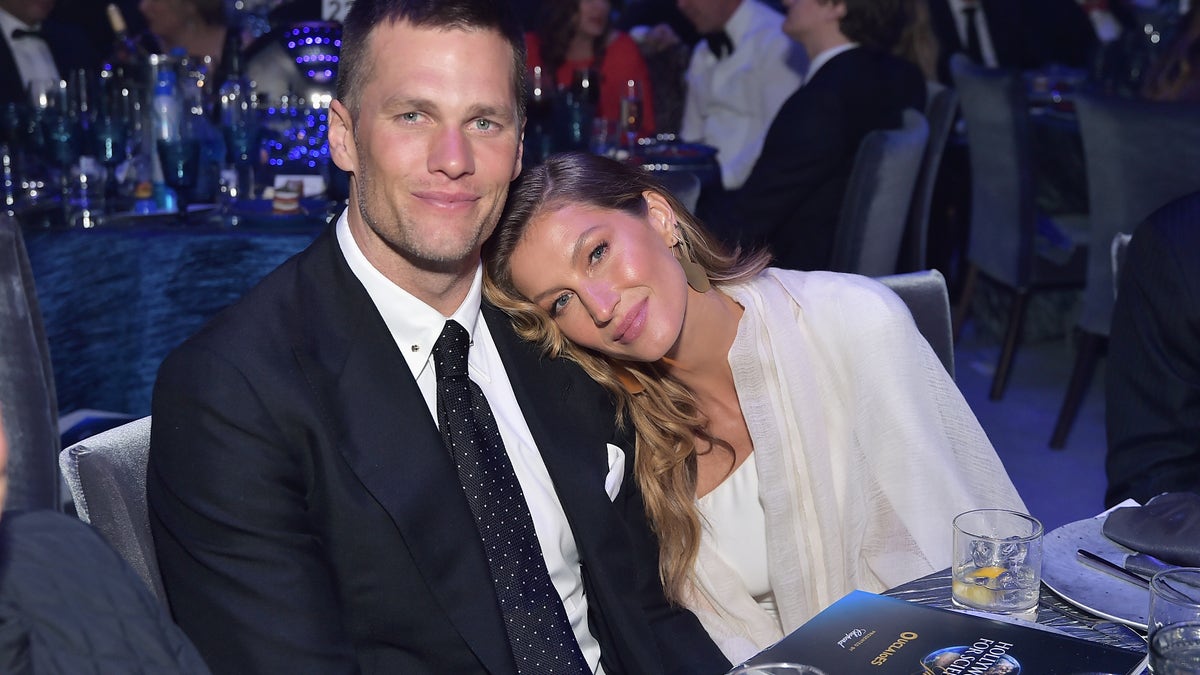 Gisele Bündchen in a white shirt and cream cardigan puts her head on Tom Brady's shoulder in a suit