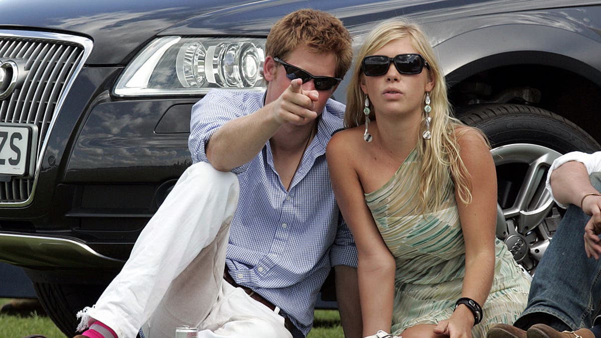 prince harry pointing at the camera as he sits next to Chelsy Davy.