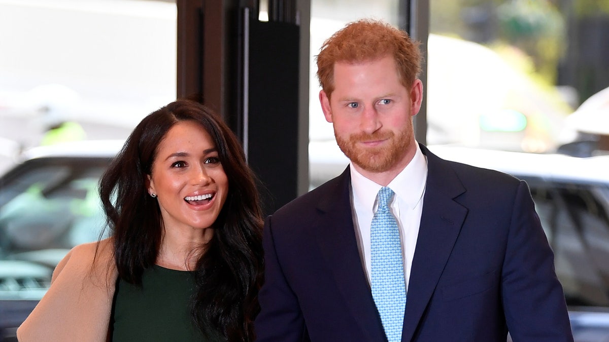 Meghan Markle in a green dress and camel colored jacket walks with Prince harry in a navy suit and teal tie