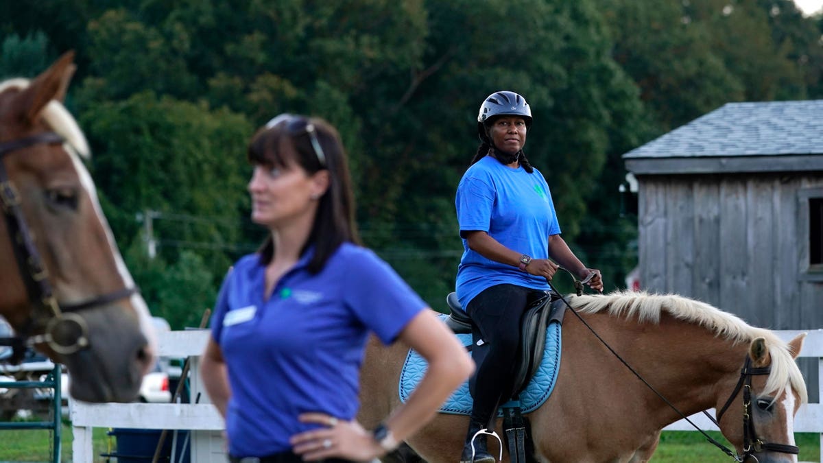 Woman standing in the foreground while a woman rides a horse in the background