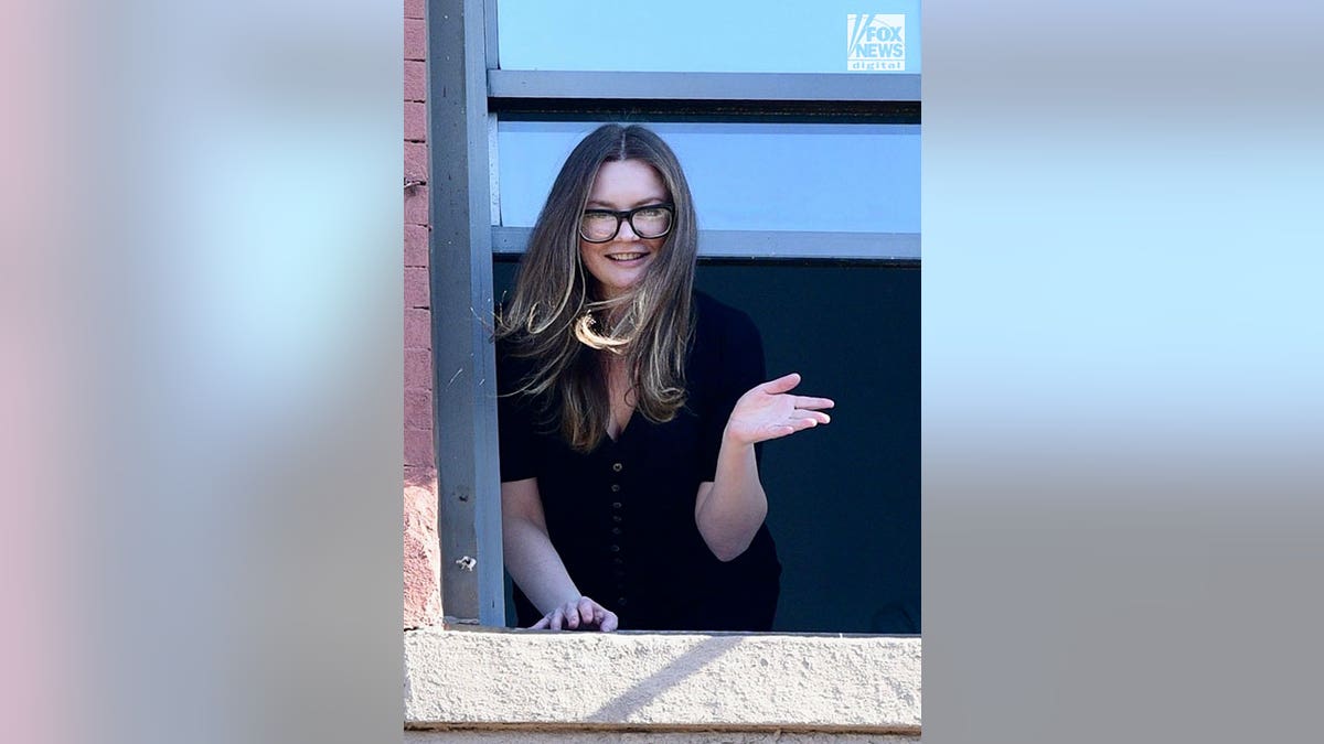 Anna Sorokin wears black top and glasses as she waves out a window of a brick building