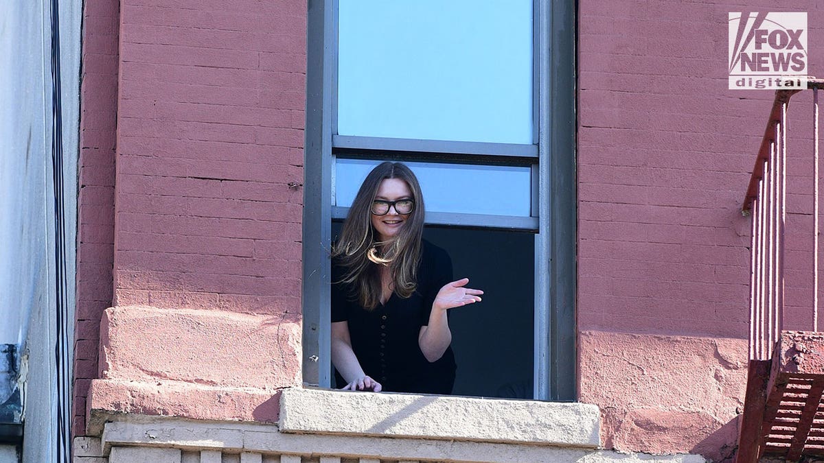 Anna Sorokin wears black top and glasses as she waves out a window of a brick building