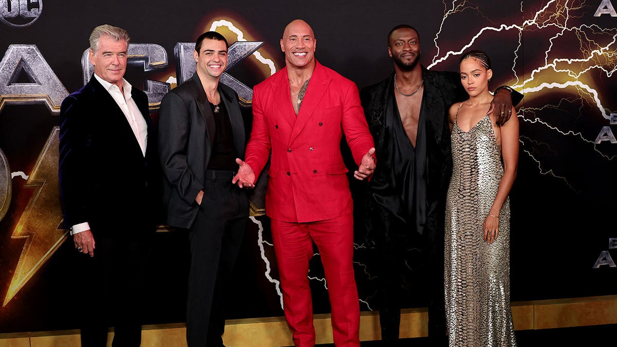 Dwayne "The Rock" Johnson smiles on the Black Adam red carpet wearing a red suit wiith costars Pierce Brosnan, Noah Centineo, Aldis Hodge and Quintessa Swindell