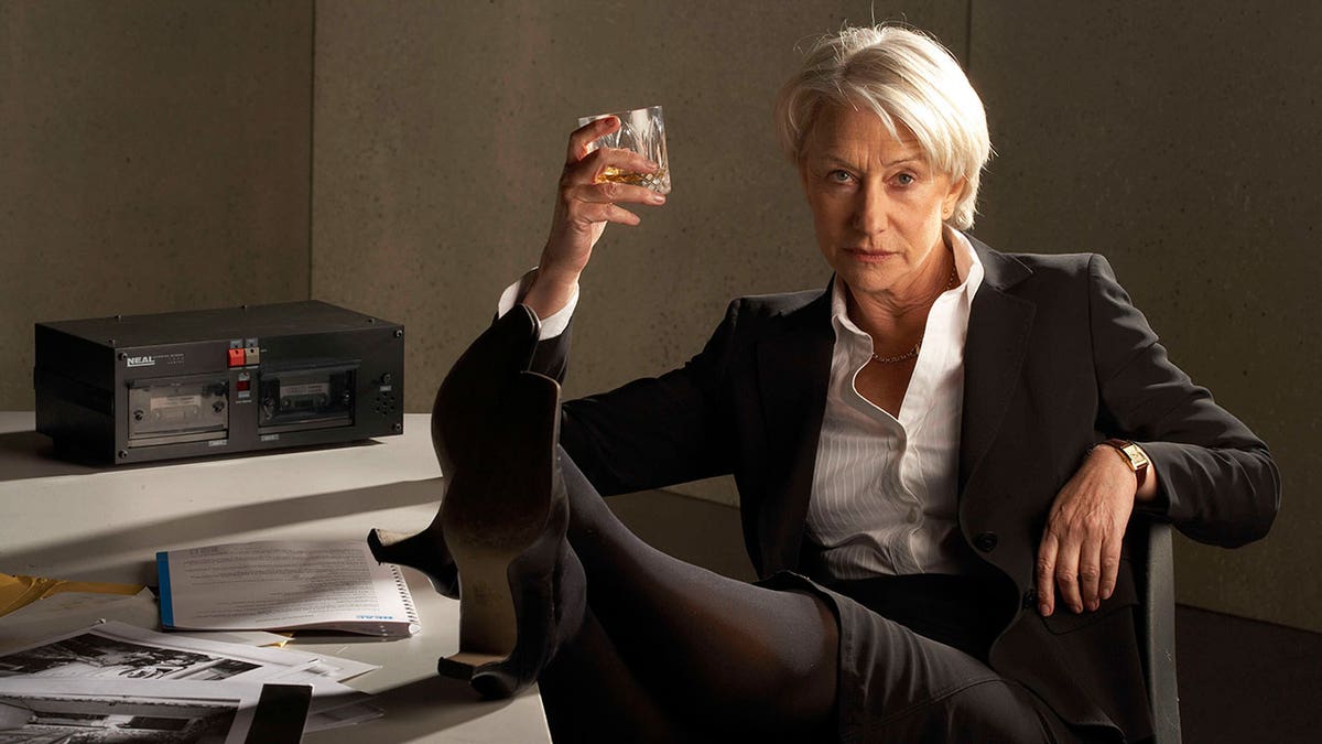 Dame Helen Mirren wears black suit and drinks scotch at her desk as a detective.