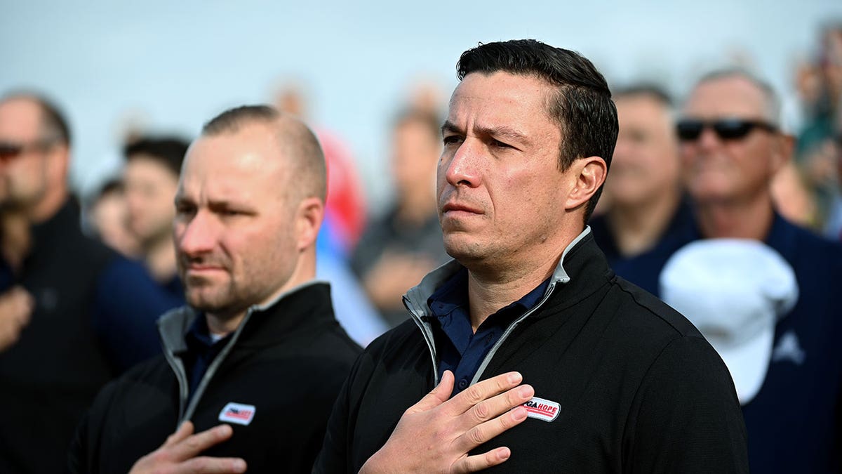 Two men stand in the foreground with their hands over their hearts during the Pledge of Allegiance