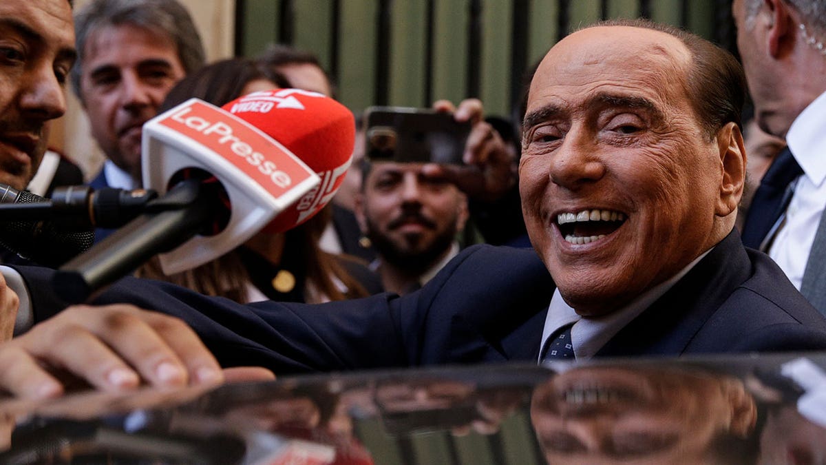 Silvio Berlusconi smiles as mics surround him and he gets into his car