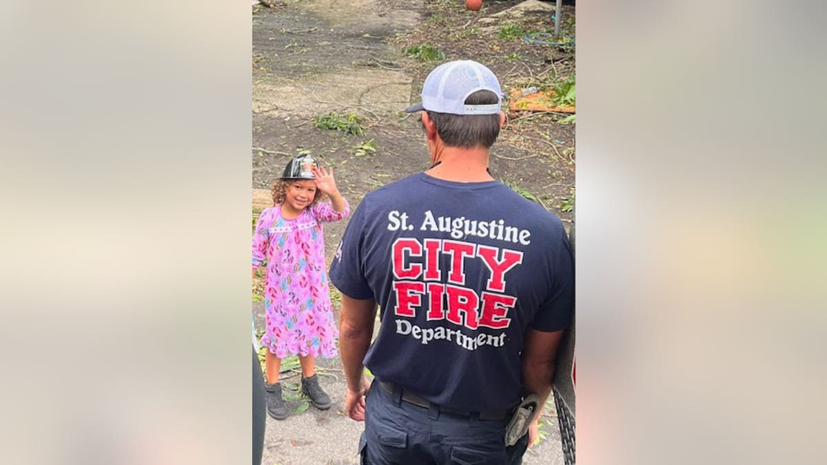 Little girl waves at the camera while wearing a firefighter hat