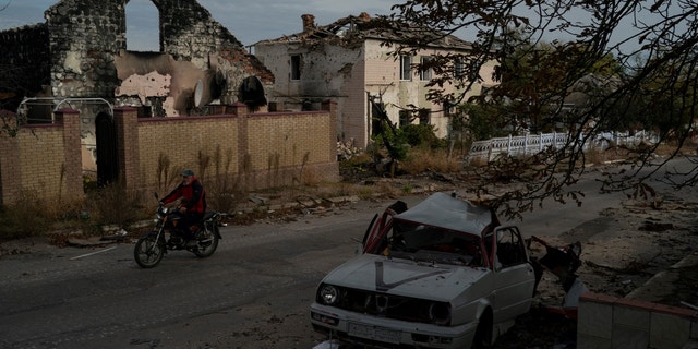 A man drives his motorcycle past a destroyed car in the retaken village of Velyka Oleksandrivka, Ukraine, Wednesday, Oct. 12, 2022.