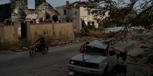 A man drives his motorcycle past a destroyed car in the retaken village of Velyka Oleksandrivka, Ukraine, Wednesday, Oct. 12, 2022. 