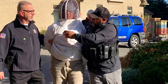 Rorie S. Woods, 55, of Hadley, Mass., center, wears a beekeeping suit while taken into custody by Hampden County Sheriff's Department officers, in Longmeadow, Mass., Wednesday, Oct. 12, 2022. 