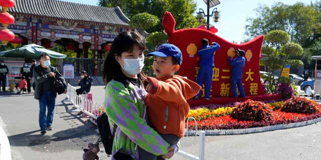 A woman wearing a mask carries a child past workers decorating an exhibition celebrating the 20th Party Congress in Beijing.