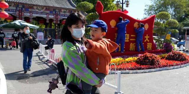 Wearing a mask, she walks past workers as they decorate a display celebrating Beijing's 20th Party Congress, holding a child.