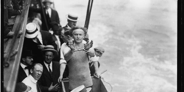 Hungarian-born American magician, escape artist and psychic debunker Harry Houdini (1874-1926) showed his handcuffs as he stood in a wooden box on a boat and prepared to be submerged, a predicament from which he would escape to the delight of onlookers, East River, July 7, 1912. 