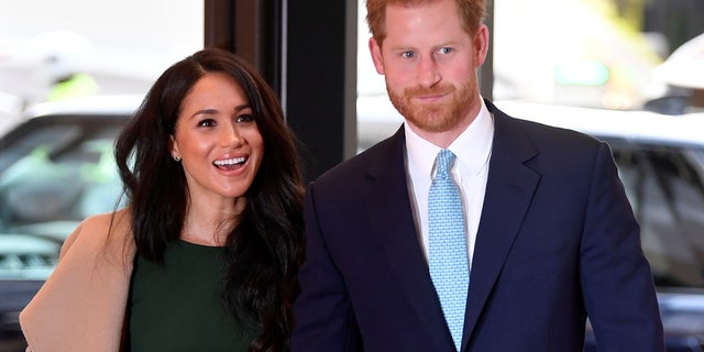 Meghan Markle in a green dress and camel colored jacket walks with Prince harry in a navy suit and teal tie