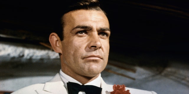 Sean Connery, arsenic James Bond, leaning against a barroom and looking retired crossed nan room. Connery is wearing a achromatic tuxedo and front necktie pinch a reddish carnation successful his lapel.