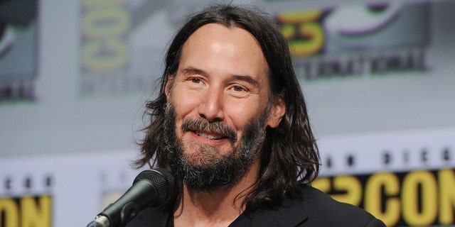 Keanu Reeves in a black shirt smiles as he speaks into a microphone at a Comic Con panel