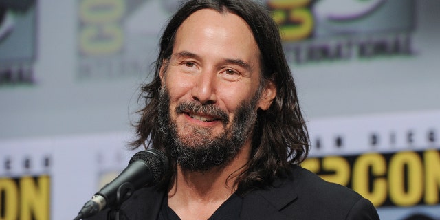 Keanu Reeves in a black shirt smiles as he speaks into a microphone at a Comic Con panel