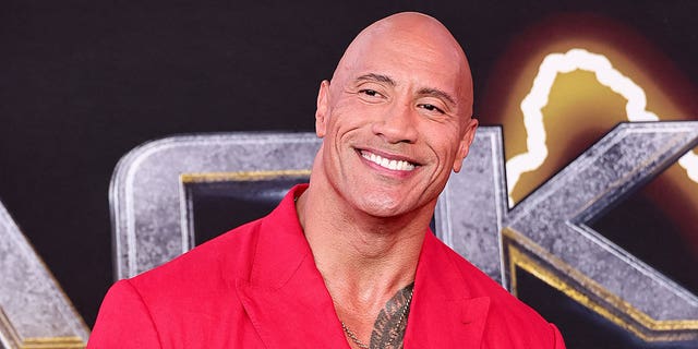Dwayne "The Rock" Johnson smiles on the Black Adam red carpet wearing a red suit