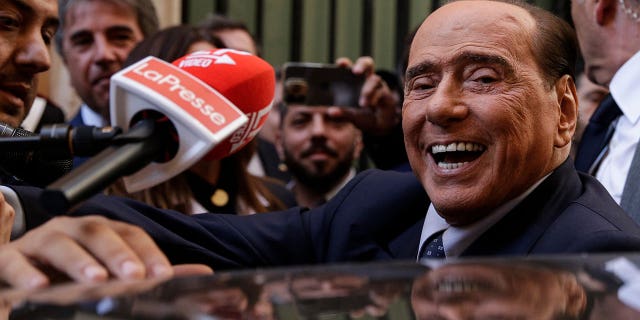 Silvio Berlusconi smiles as mics surround him and he gets into his car