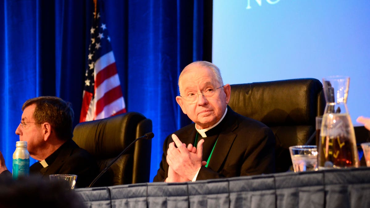 Catholic Archbishop sitting at a table, on stage, with his hands together.