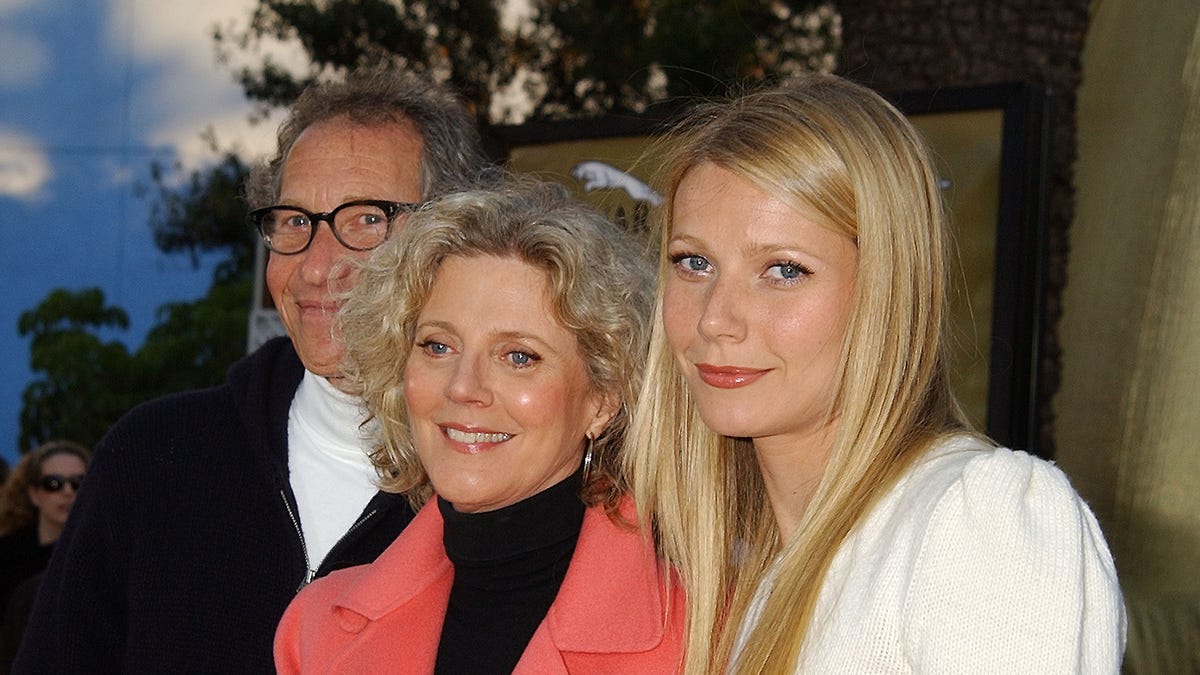 Bruce Paltrow smiles with his wife Blythe Danner in a coral suit jacket and black top and daughter Gwyneth in a white top on the red carpet