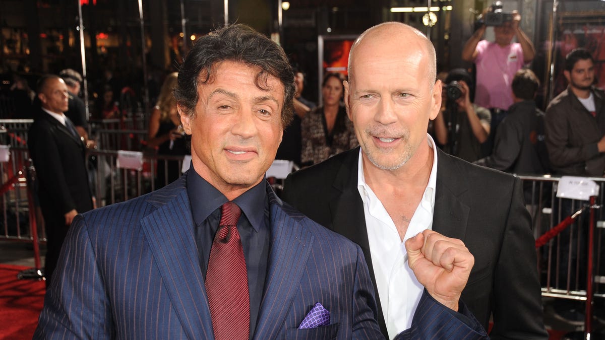 Sylvester Stallone in a blue suit and red tie on the red carpet with Bruce Willis in a white shirt and black ssuit