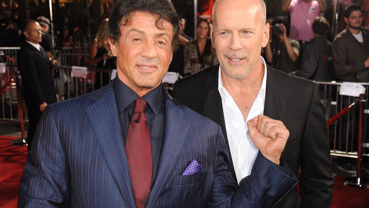 Sylvester Stallone in a blue suit and red tie on the red carpet with Bruce Willis in a white shirt and black ssuit