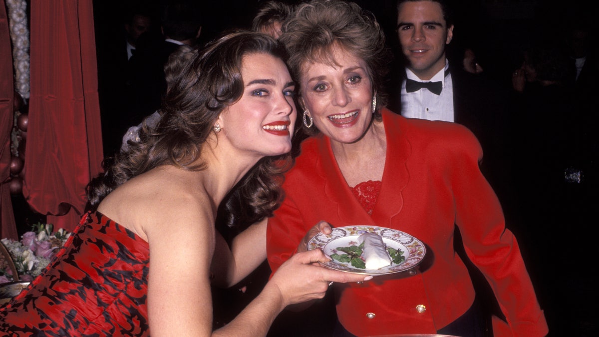 Brooke Shields in a red dress smiles and holds a plate with Barbara Walters in a red suit at the Plaza Hotel in 1991