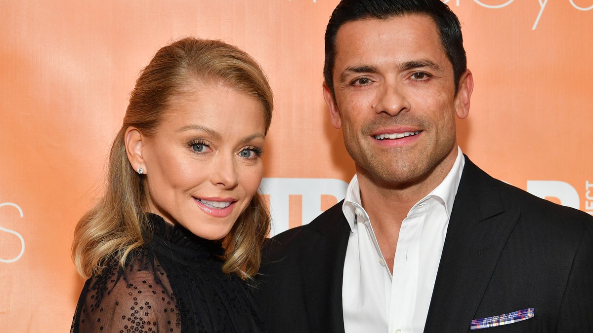 Kelly Ripa smiles in a black dress next to her husband Mark Consuelos in a white button down and black suit