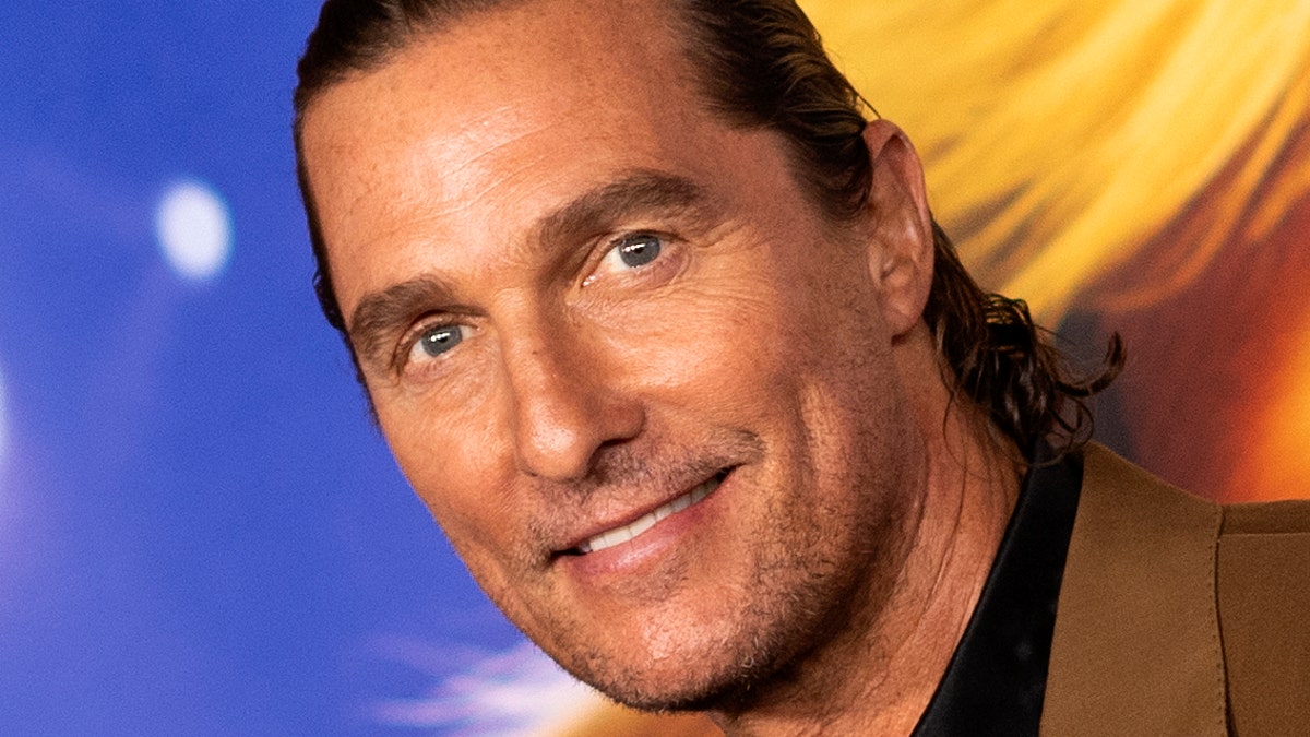 Matthew McConaughey smiles in a black shirt and brown jacket on the red carpet